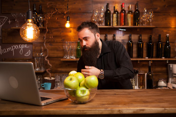 Bearded young bartender checking himself in the laptop.