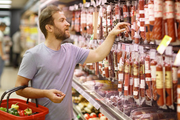 Customer standing in meat section and choosing salami or sausages. Bearded man holding basket and...