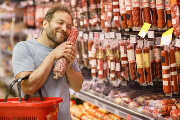 Happy man holding in hands near face salami and smiling. Bearded customer standing with closed eyes...