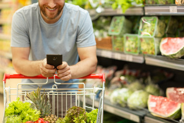 Unrecognizable man with beard holding phone in hands. Male customer leaning on trolley with vegetables and fruit. Shelves with variety of products in supermarket on background.