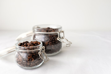 Obraz na płótnie Canvas Dark roasted coffee beans in small and large glass jar on white cotton fabric background in natural light with empty text copy space on white wall background