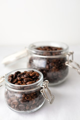 Dark roasted coffee beans in small and large glass jar on white cotton fabric background in natural light with empty text copy space on white wall background