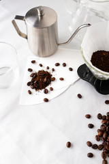 Coffee making equipment on white table top and white wall background. Roasted coffee beans in glass jar, glass jug and dipper, stainless pitcher, paper filter, ground coffee