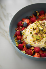 Beeren Obstsalat mit Erdbeeren, Himbeeren, Blaubeeren, Pistazien, Zitronen Zesten, Leinöl Kurkuma Quark Creme in blauer Schale als Frühstück auf Marmor Hintergrund
