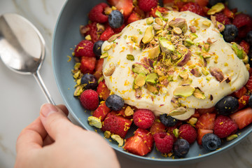 Frau isst Beeren Obstsalat mit Erdbeeren, Himbeeren, Blaubeeren, Pistazien, Zitronen Zesten, Leinöl Kurkuma Quark Creme in blauer Schale als Frühstück auf Marmor Hintergrund