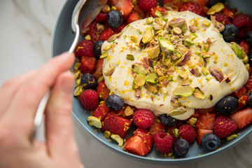 Frau isst Beeren Obstsalat mit Erdbeeren, Himbeeren, Blaubeeren, Pistazien, Zitronen Zesten, Leinöl Kurkuma Quark Creme in blauer Schale als Frühstück auf Marmor Hintergrund