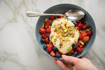 Frau isst Beeren Obstsalat mit Erdbeeren, Himbeeren, Blaubeeren, Pistazien, Zitronen Zesten, Leinöl Kurkuma Quark Creme in blauer Schale als Frühstück auf Marmor Hintergrund