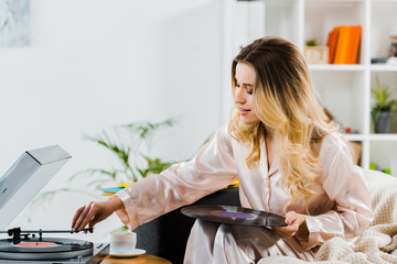 Curly woman in pyjamas holding vinyl record and using gramophone