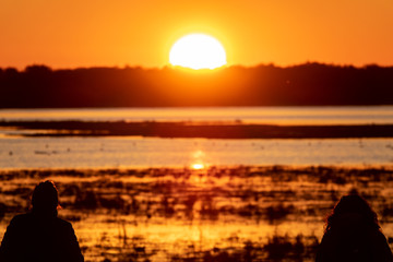 Naklejka na ściany i meble Two woman silhouettes observing sunrise over lake