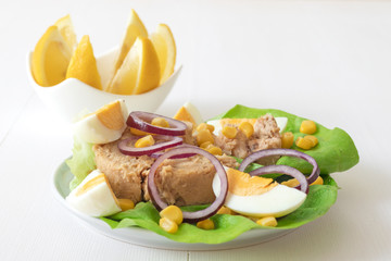 Canned tuna with eggs, suggar corn, red onion rings on green lettuce leaf, slice of lemon in pocelain bowl..Tuna fish salad. White wooden table background.