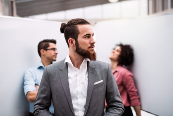 A young businessman in suit standing in office, colleagues in the background.