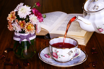 Tea break time. Tea cup, tea pot, wildflowers in vase and open book on wooden background. Teapot...