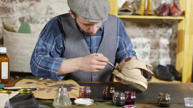 Male shoemaker in flat cap and vest sitting at desk in his workshop and applying leather dye to shoe with brush