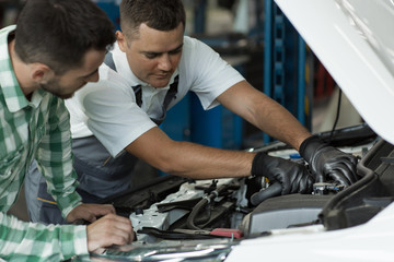 Fototapeta na wymiar Qualified mechanic explaining client breakage in car at service. Repair man in white shirt and uniform showing problem in hood of vehicle and owner of automobile listening carefully.