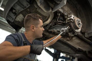 Bottom view of muscular mechanic standing under car, keeping crowbar and fixing running gear of automobile at service. Qualified repairman in uniform and protective gloves in process of fixing car.