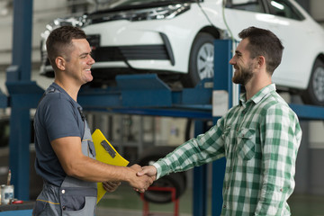 Smiling worker of car service in uniform and satisfied client shaking hands. Happy man in checkered...
