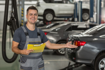 Portrait of positive repairman in uniform showing with hand auto service equipped with modern technology. Smiling mechanic keeping folder, gesturing with hand on cars and looking at camera.