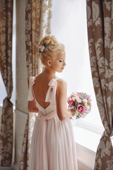 beautiful blonde young woman stands in the image of a bride with a wedding bouquet of roses and orchids standing near the window in a dress with a naked back.