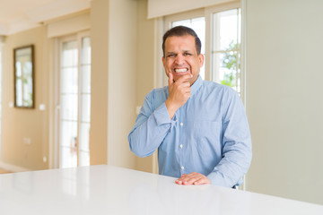 Middle age man sitting at home looking confident at the camera with smile with crossed arms and hand raised on chin. Thinking positive.