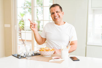 Middle age man eating asian food with chopsticks at home with a big smile on face, pointing with hand and finger to the side looking at the camera.