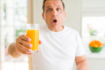 Middle age man drinking a glass of orange juice at home scared in shock with a surprise face, afraid and excited with fear expression