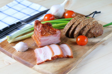 A piece of bacon on a chopping board with vegetables and bread.