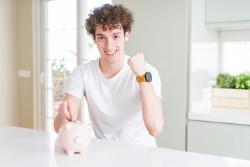 Young man investing money using piggy bank at home screaming proud and celebrating victory and success very excited, cheering emotion