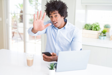 African American business man using smartphone and laptop with open hand doing stop sign with serious and confident expression, defense gesture