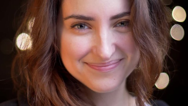 Close-up portrait of young caucasian girl smiling modestly into camera on blurred lights background.