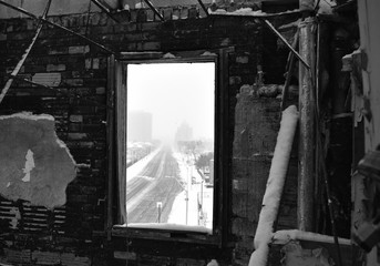 Looking through the broken window of abandoned brick building of a street in the city during a snowstorm in monochrome.