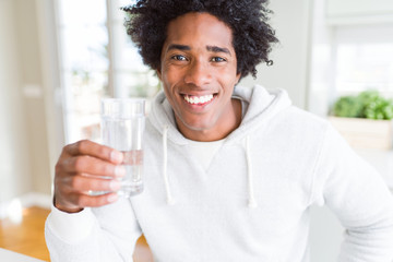 African American man drinking a glass of water at home with a happy face standing and smiling with...