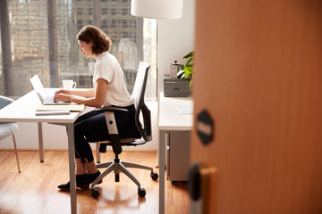 Businesswoman Sitting At Desk Working On Laptop In Modern Office Viewed Through Door Way - Powered by Adobe