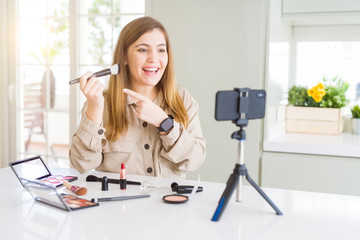 Beautiful young influencer woman recording make up video tutorial very happy pointing with hand and finger