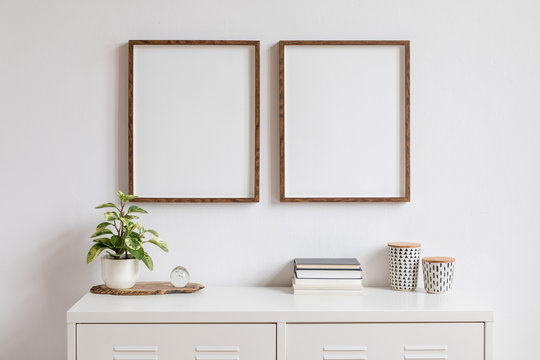 Minimalistic home decor of interior with two brown wooden mock up photo frames on the white shelf with books, beautiful plant in stylish pot and home accessories. White wall. 