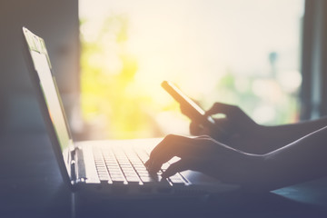 Technology business and working concept. Close up woman hand using keyboard laptop and smart phone with mirror sun light abstract background.