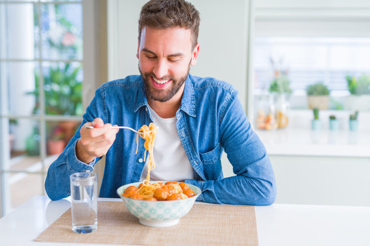Handsome man eating pasta with meatballs and tomato sauce at home while smiling at the camera