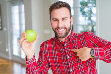 Handsome man eating fresh healthy green apple with surprise face pointing finger to himself