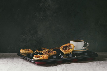 Traditional Portuguese egg tart dessert Pasteis Pastel de nata in black baking tray with cup of black coffee standing on linen table cloth near dark grey wall.