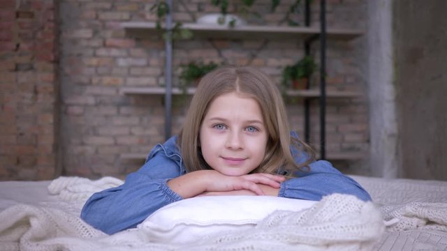 happy childhood, portrait of beautiful little girl with blue eyes lying on pillow on bed in room at home