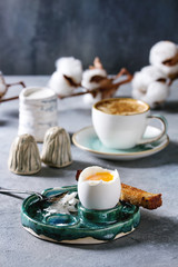 Breakfast with cup of coffee and soft boiled egg, served in green ceramic egg cup with salt, pepper and toasted bread, jug of cream and cotton flowers over grey blue table.