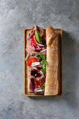 Beef baguette sandwich with tomatoes, basil, red onion served on wooden serving board over grey texture background. Flat lay, space