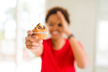 Young african american woman eating chocolate chips muffins with happy face smiling doing ok sign with hand on eye looking through fingers