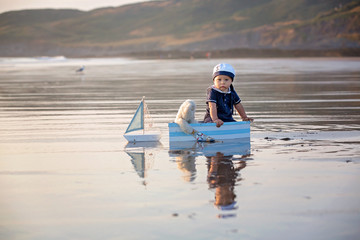 Cute baby child, sweet boy, playing with boat, teddy bear and fishes on sunset at the edge of the ocean