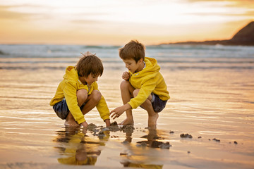 Two beautiful children, boy brothers, playing on the beach with sand and running in the water on sunset - Powered by Adobe