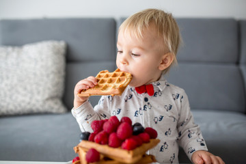 Sweet toddler birthday boy, eating belgian waffle with raspberries, blueberries, cocnut and...