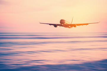 Naklejka na ściany i meble Airplane flying over blur tropical beach with smooth wave and sunset sky abstract background.