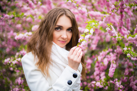 Portrait of a beautiful fair-haired girl in a white coat. The girl holds a branch of pink cherry blossoms. Cherry blossom. Horizontal photography
