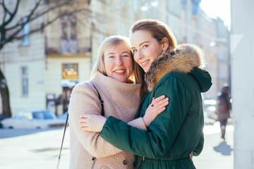 Portrait of blond caucasian mid-adult daughter and mature mother hugging with foreheads together looking at camera in cold weather in city street on background. Bonding, connect, family concept.