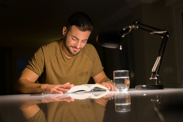 Young handsome man studying at home, reading a book at night
