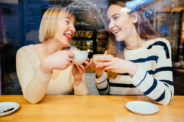 View through the window of young beautiful blonde girl talking to her mom an interesting secret, they sit at cafe, happy and laugh. Bonding, different generationsconnect, family concept.